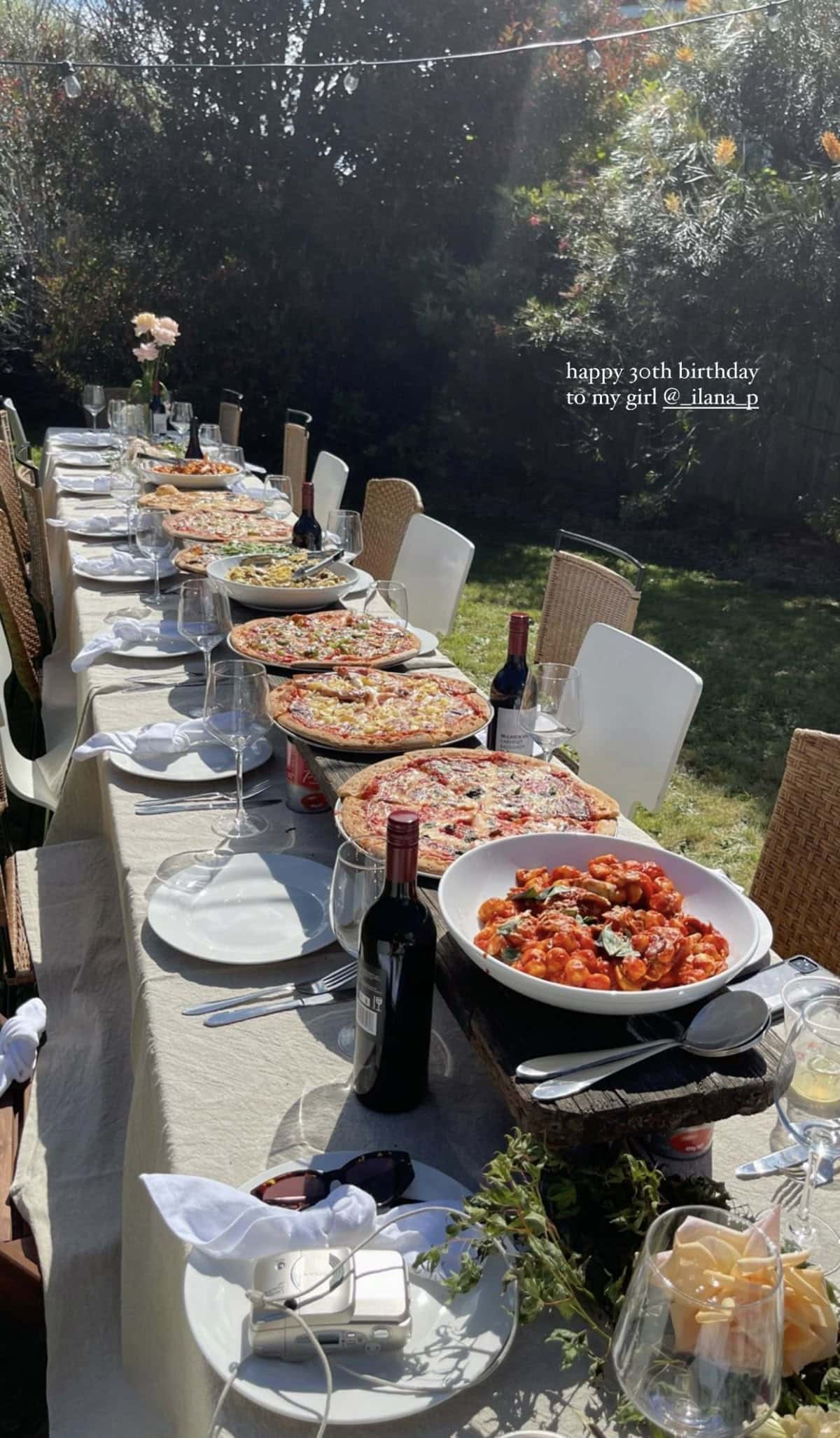 Long outdoor birthday table with pizzas and red wine bottles down the centre