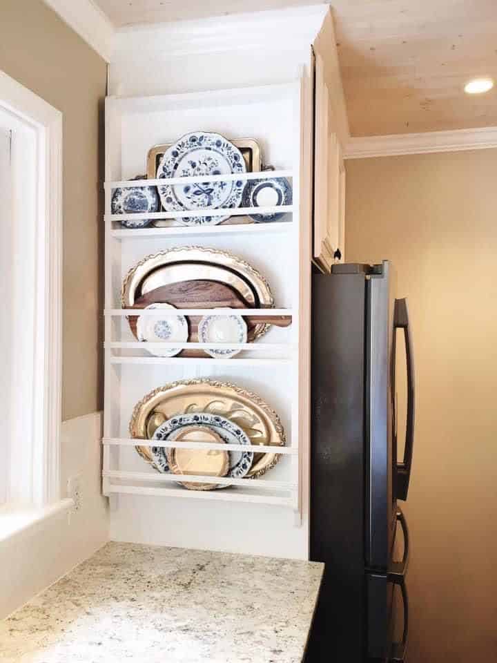 Narrow white plate rack beside the fridge layered with oversized platters and silver trays