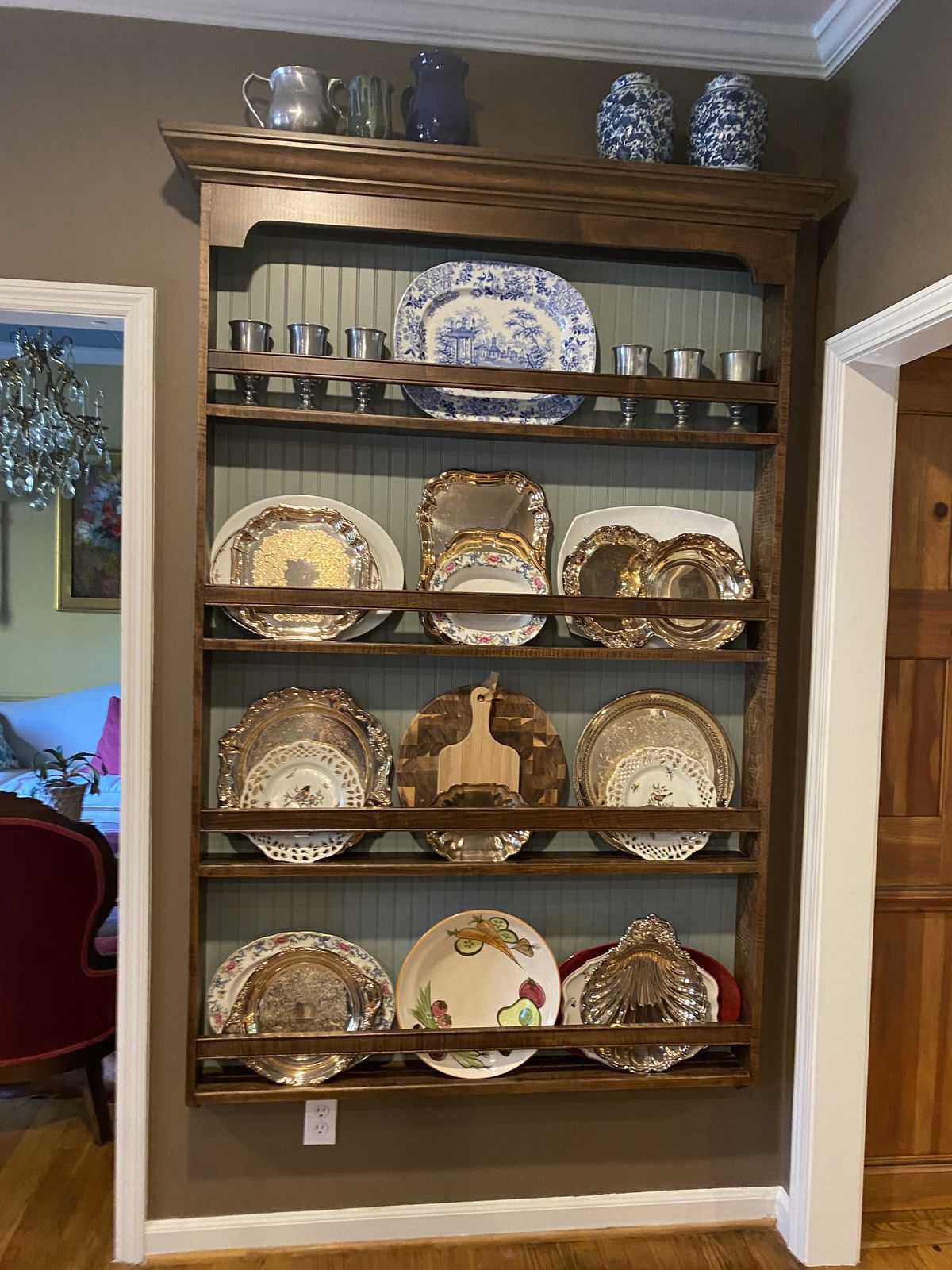 Dark wood plate rack wall displaying silver trays, pewter goblets, and vintage plates
