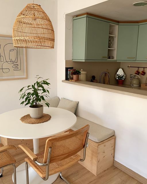 Corner built-in wooden bench with a round white pedestal table and cane chairs beside a sage green kitchen pass-through