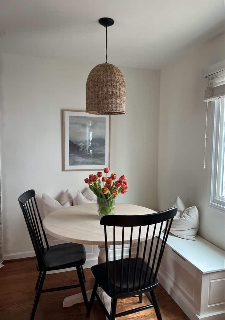 Round whitewashed pedestal table with black spindle chairs and a white storage bench under a woven dome pendant