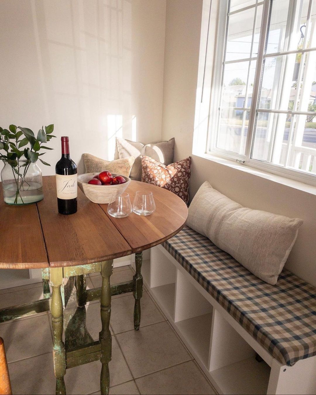 White cube-storage bench with a checked cushion and floral pillows beside a drop-leaf table with green painted legs