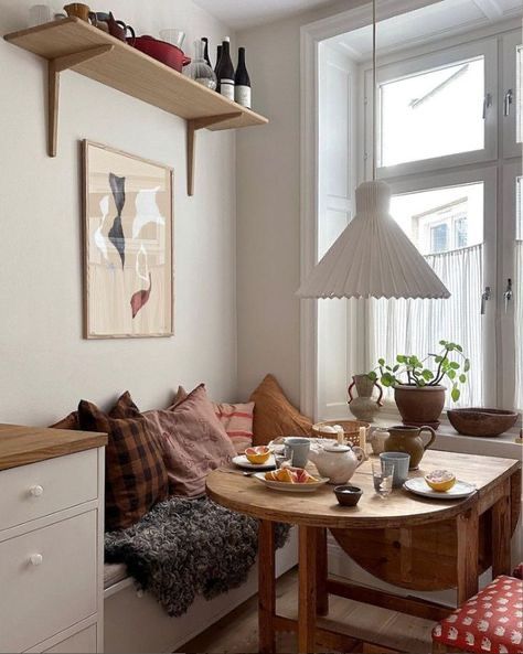 Compact white built-in bench layered with warm-toned cushions beside a wooden drop-leaf table