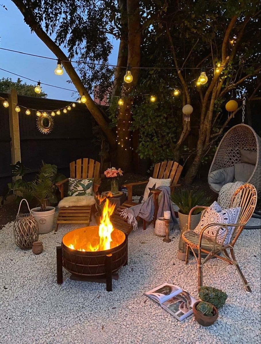 Cosy backyard fire pit lounge on white gravel with string lights, timber Adirondack chairs and a hanging egg chair at dusk