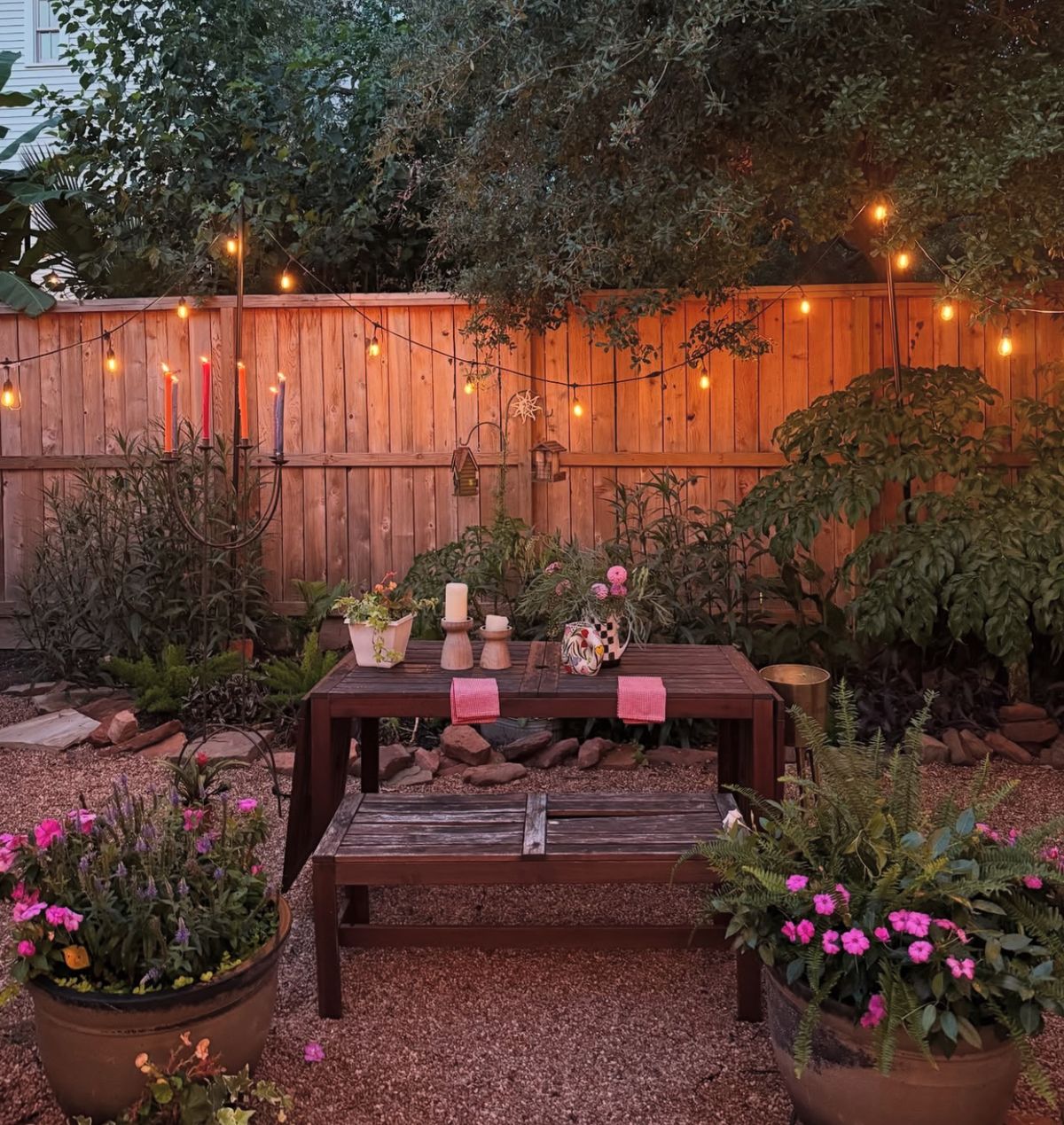 Candlelit outdoor dinner table with a taper candelabra, pillar candles and pots of pink flowers by a fence