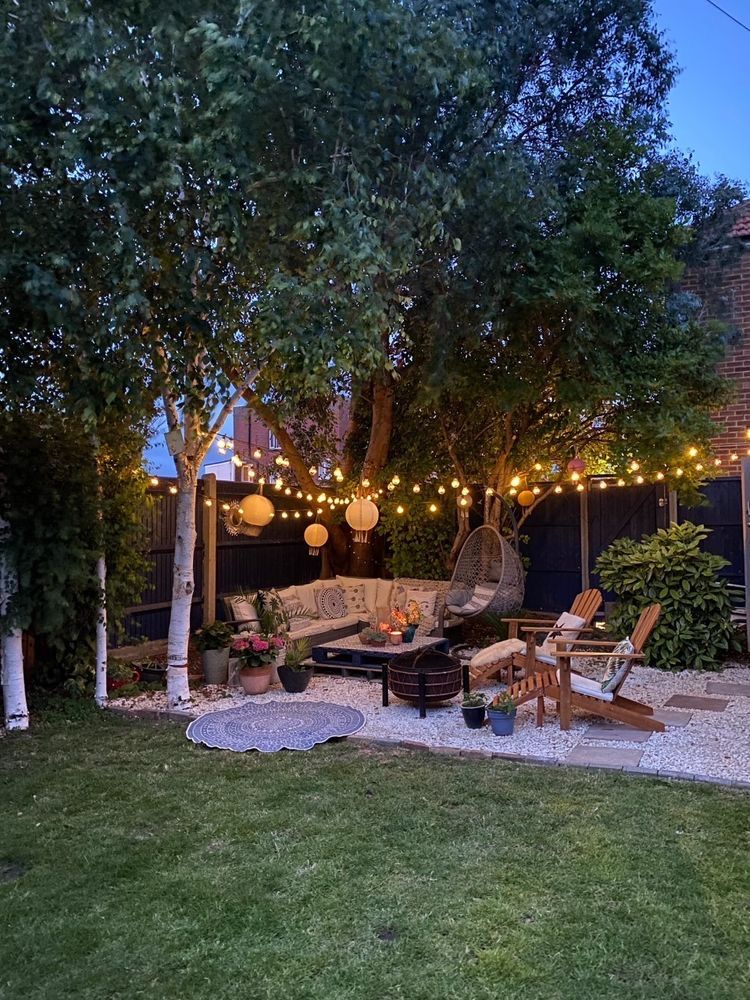 Wide view of a gravel lounge area meeting the lawn under mature trees, with string lights and a fire pit