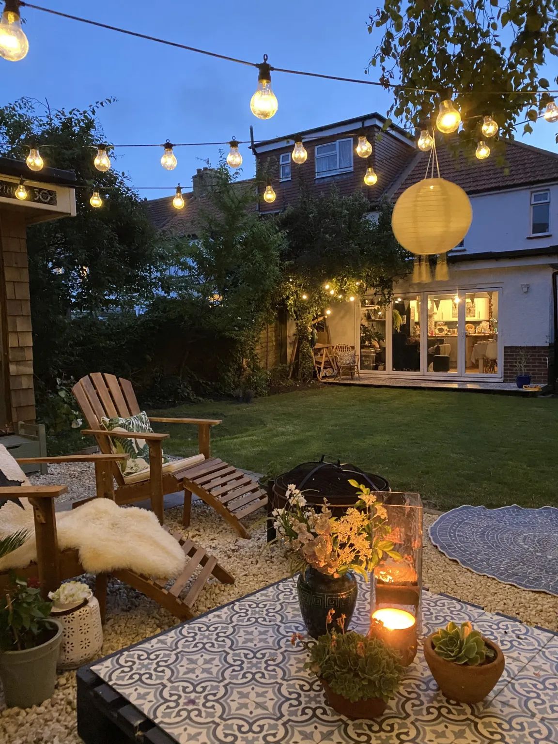 Slatted timber recliner loungers draped with sheepskins beside a tiled table at the edge of a lawn