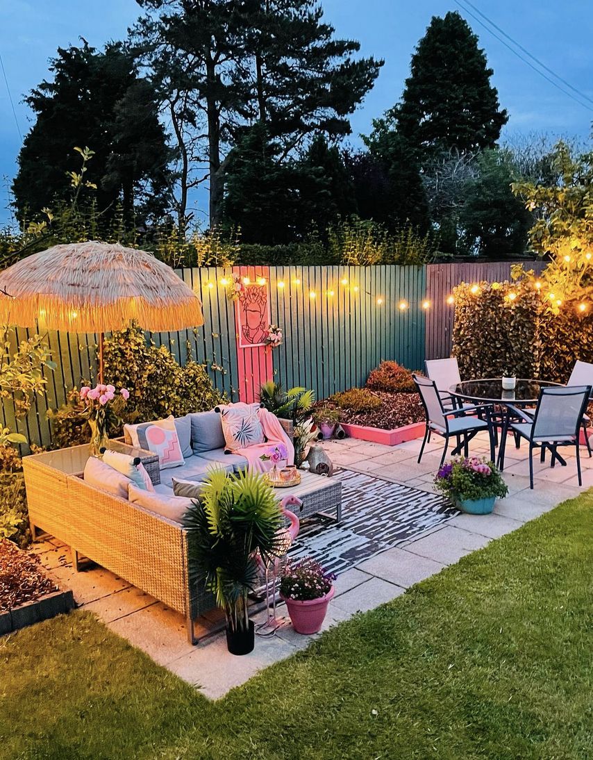 Colourful boho garden lounge at dusk with a raffia parasol, rattan corner sofa, painted fence, festoon lights and pink accents