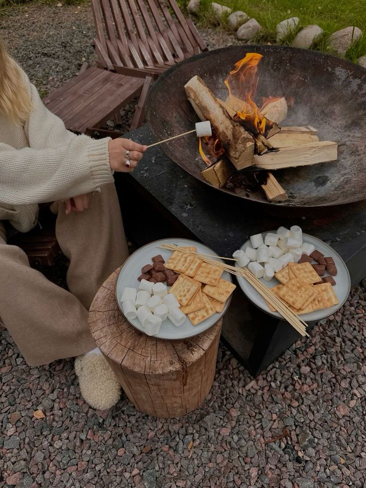 S'mores station by a cast iron fire bowl with a stump table holding marshmallows, crackers and chocolate