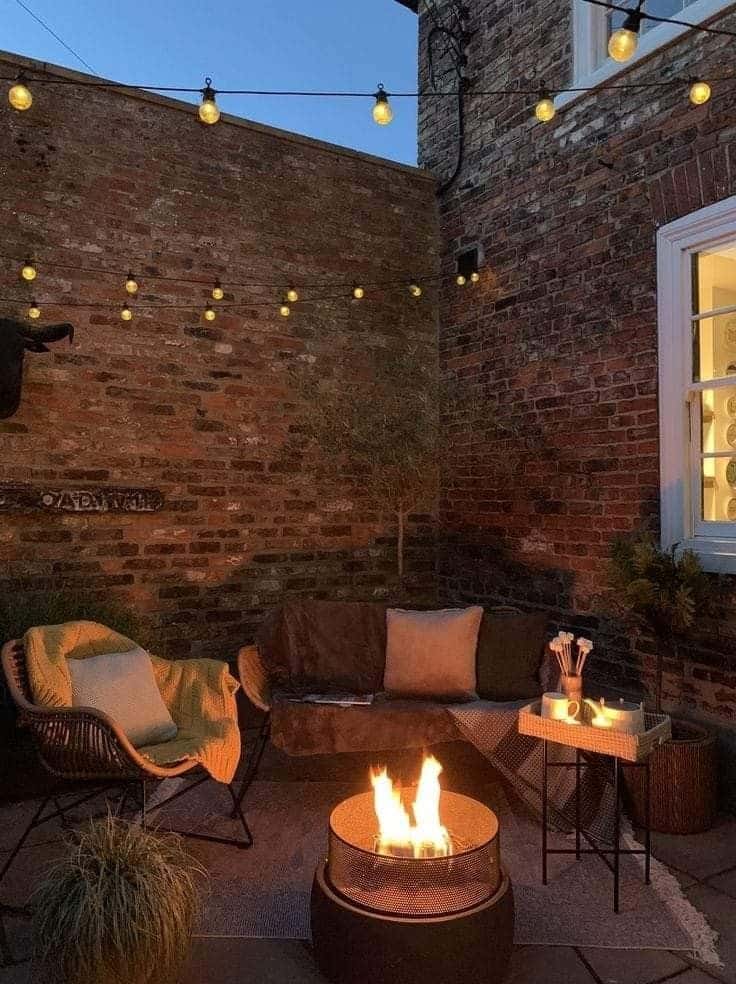 Walled brick courtyard at dusk with a modern fire pit sofa and festoon lights strung overhead