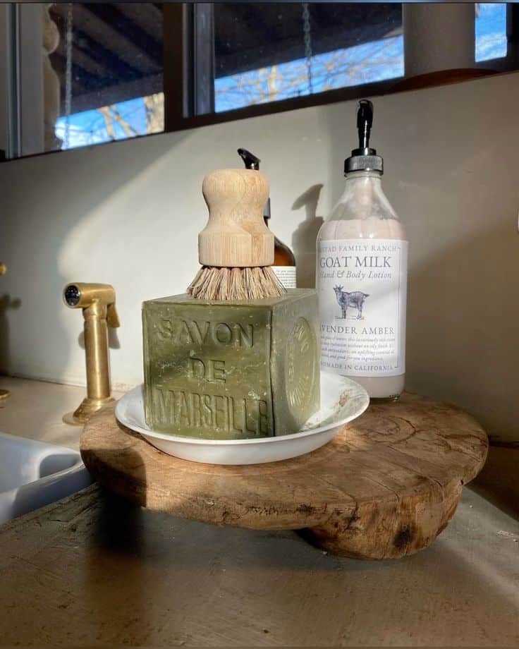 Rustic wooden round holding a green Savon de Marseille soap cube, a bamboo brush and a bottle of lotion