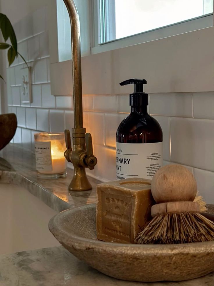 Shallow stone bowl holding a block of soap and a wooden brush beside an amber pump bottle and a candle