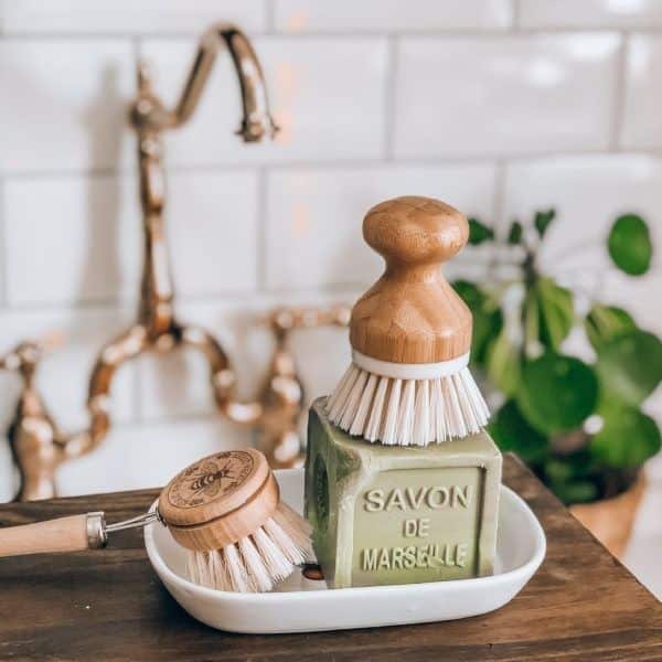 Simple white ceramic dish holding two scrub brushes and a green bar of soap on a wooden bench