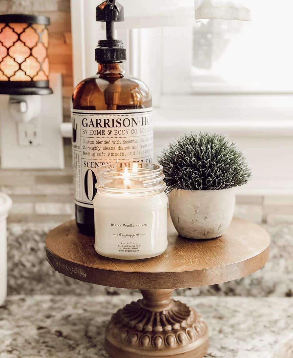 Wooden pedestal cake stand styled with an amber dish soap bottle, a lit candle and a small potted plant