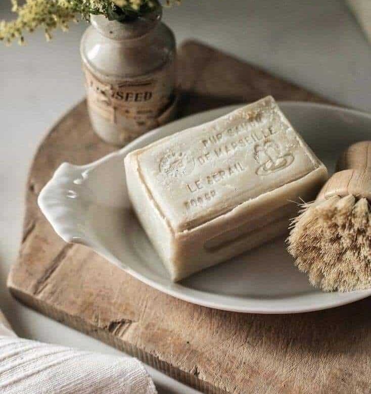 White ceramic plate on a wooden board holding a bar of Marseille soap and a natural dish brush