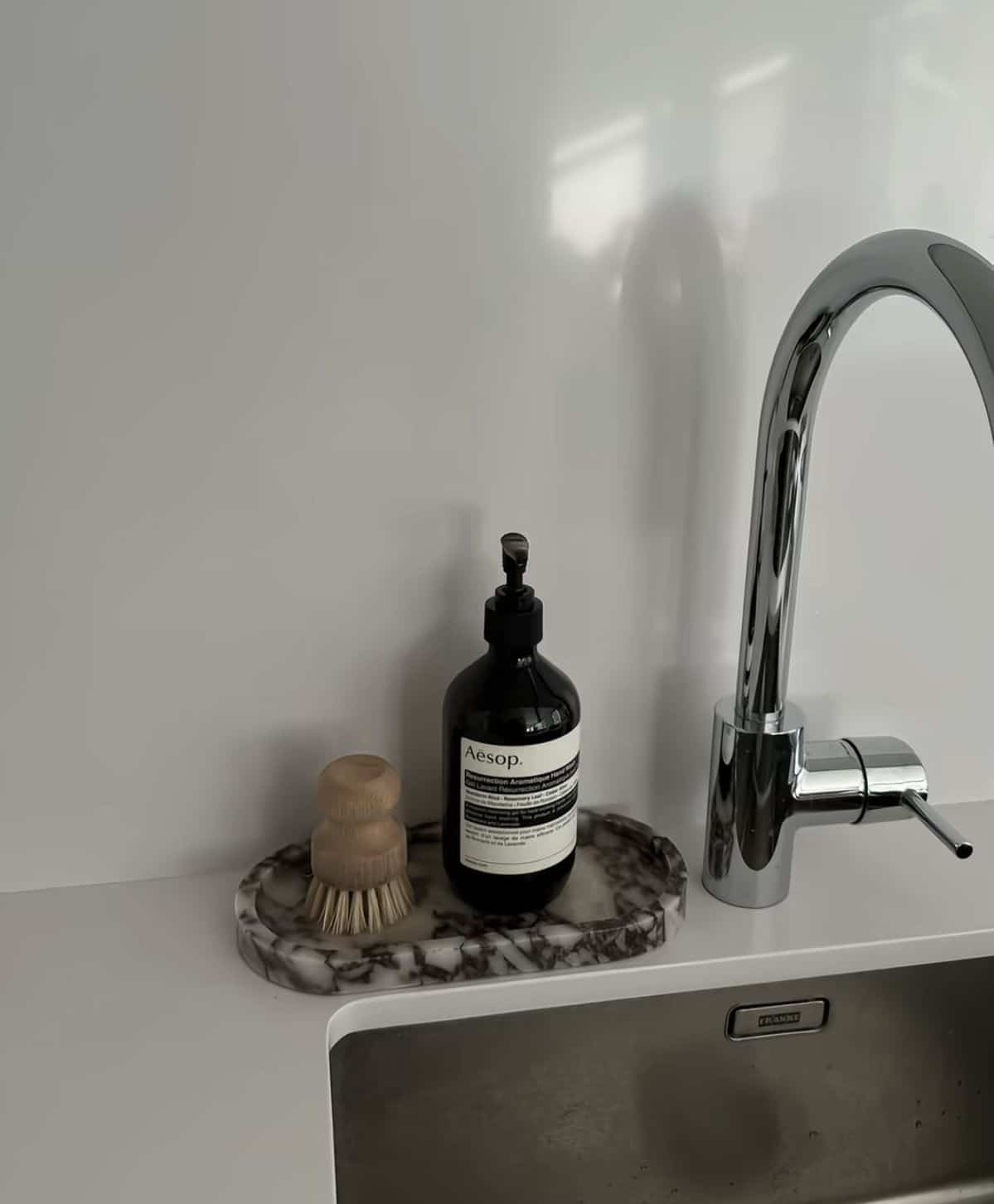 Sleek marble oval tray holding a dark soap bottle and a wooden dish brush beside a modern chrome tap