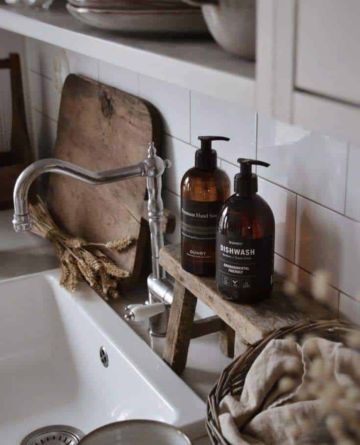 Small wooden stool used as a riser holding two amber soap bottles over a white farmhouse sink