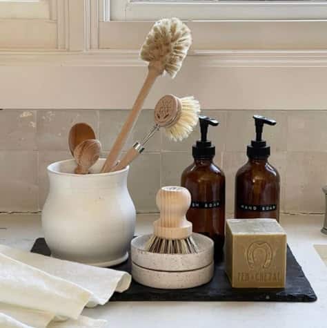 White crock of brushes and a stone double dish with a round scrubber and amber soap bottles on a slate tray