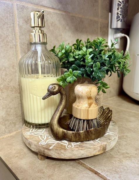 Brass swan shaped dish holding a scrub brush beside a ribbed glass soap dispenser on a round wooden riser