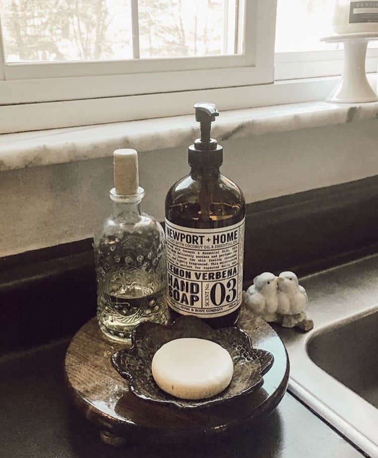 Dark glazed dish holding a white bar of soap beside an amber hand soap bottle on a wooden round