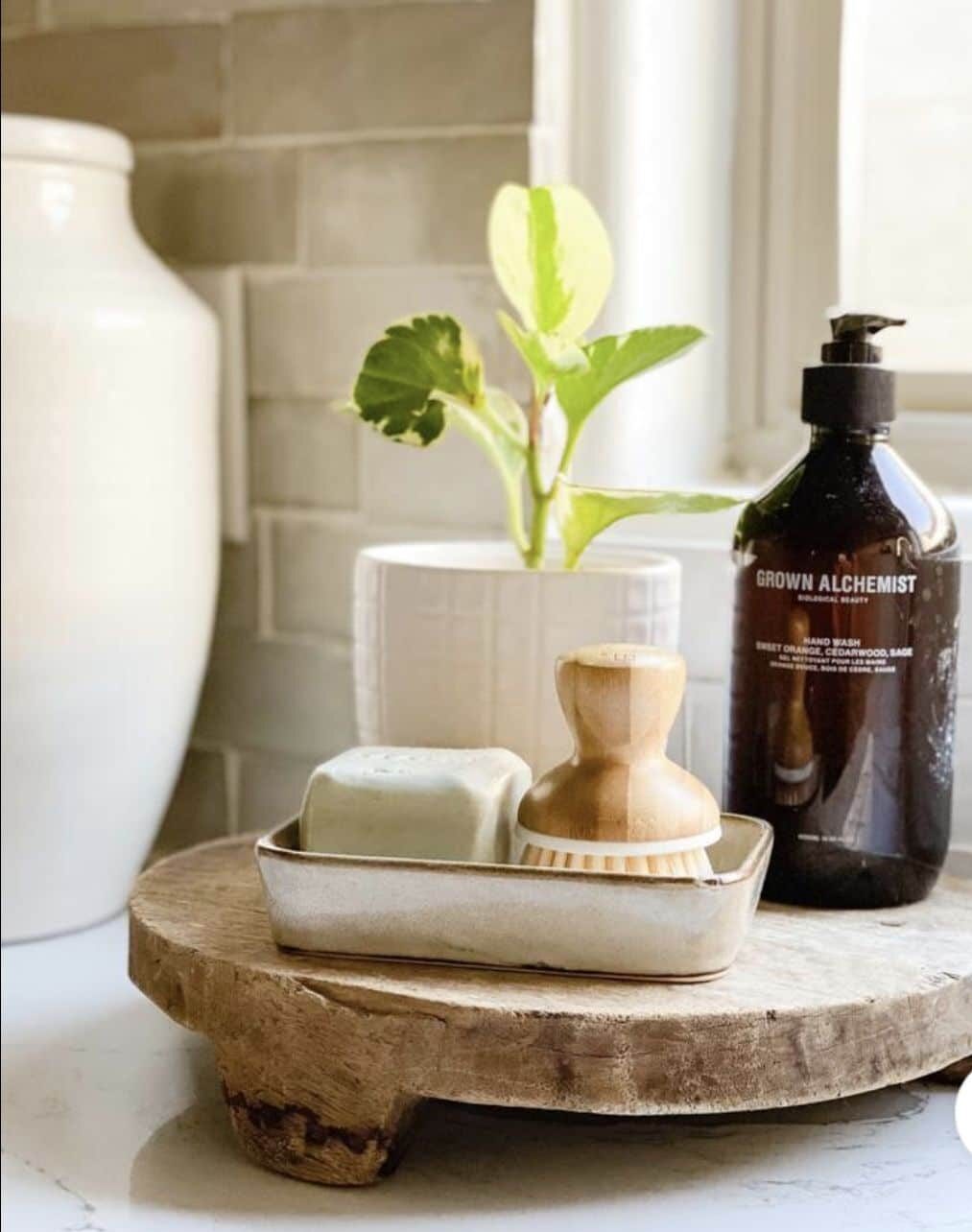 
Chunky raw wood round riser holding a small ceramic tray with a soap bar and a wooden dish brush, an amber hand wash bottle and a potted plant against sage tile
