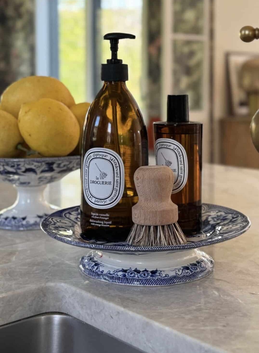 Blue and white china footed stand holding amber bottles and a wooden dish brush beside a bowl of lemons