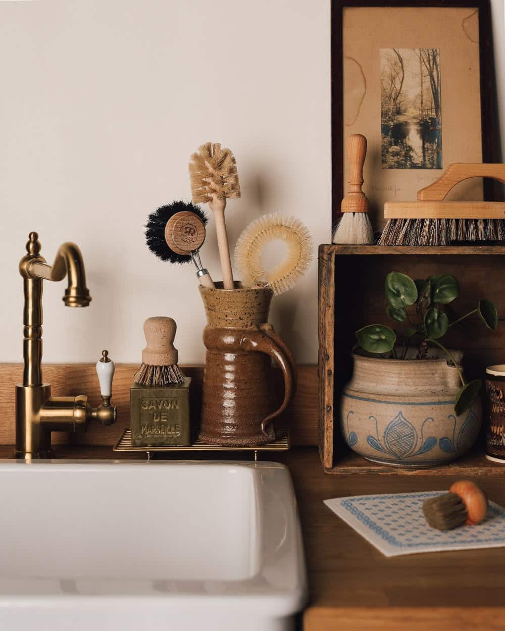 Ceramic jug filled with dish brushes on a brass tray next to a Savon de Marseille soap block by a farmhouse sink