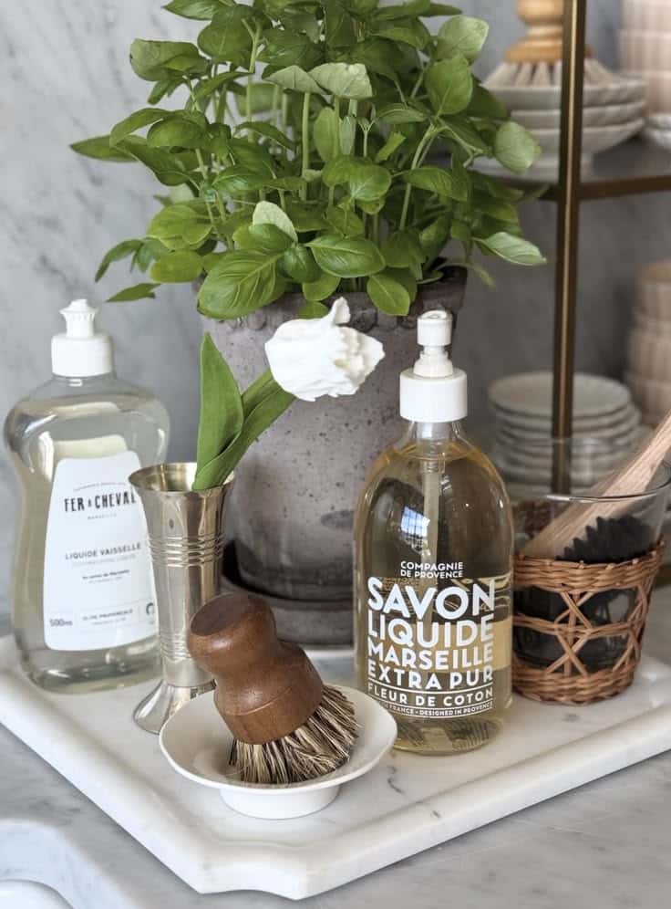 Marble board styled with dish soap bottles, a potted basil plant and a wooden dish brush