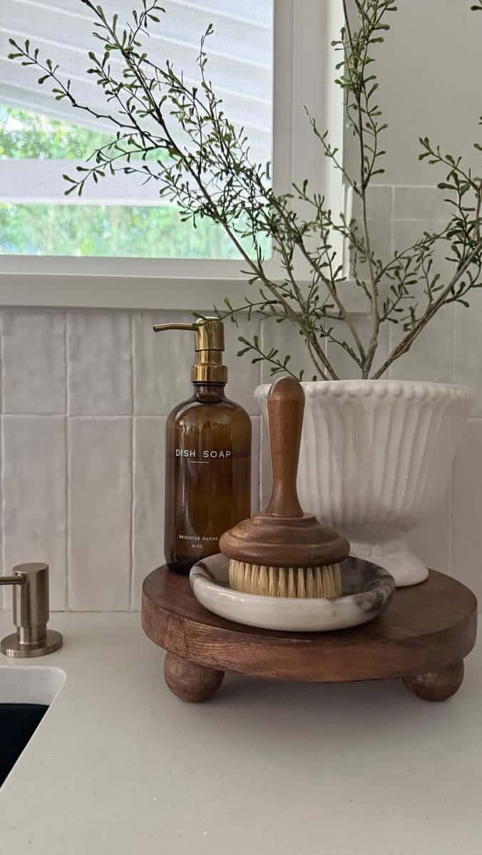Raised wooden pedestal with a marble dish, wooden brush and amber dish soap bottle beside a white urn of greenery