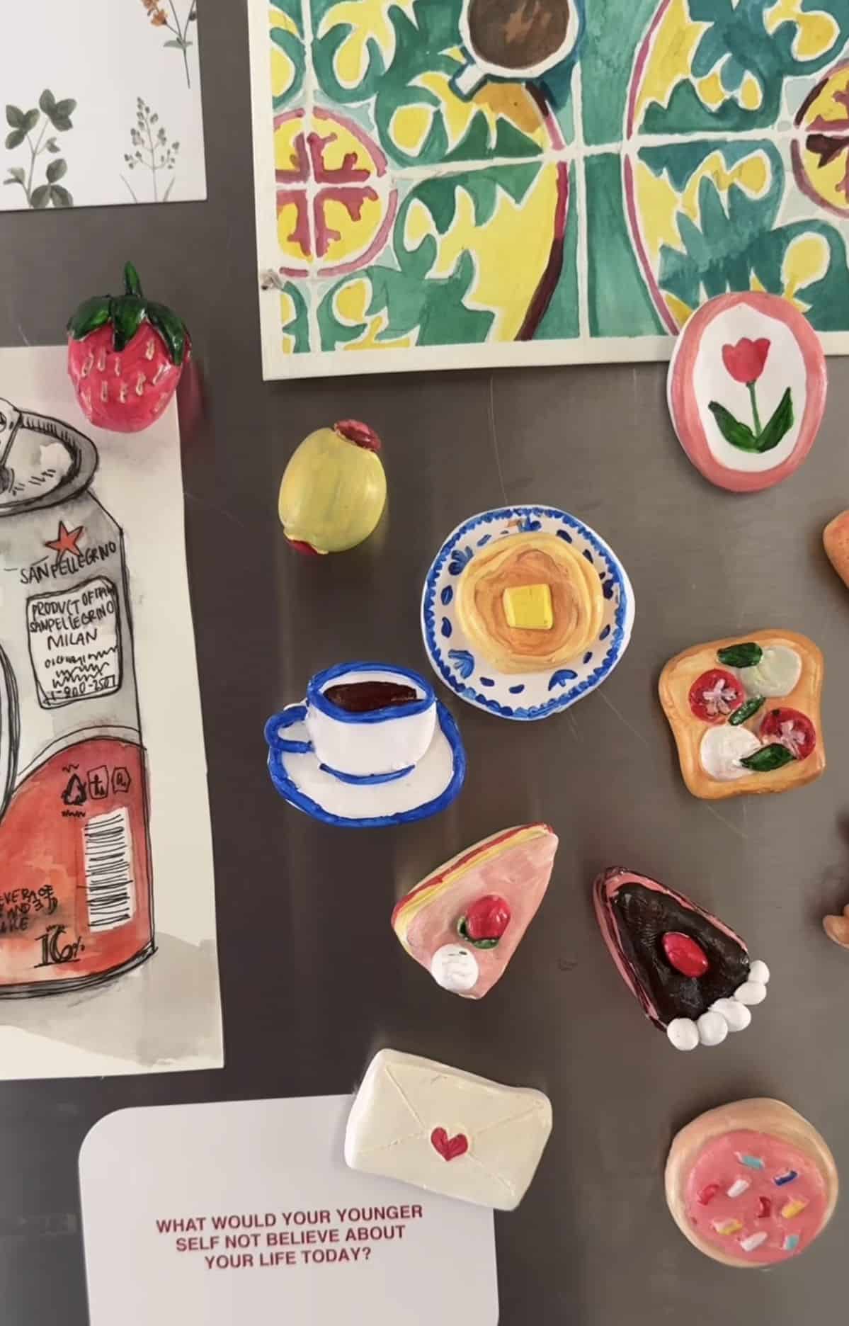 Painterly clay cafe magnets including an espresso cup, pancakes and cake slices on a dark fridge
