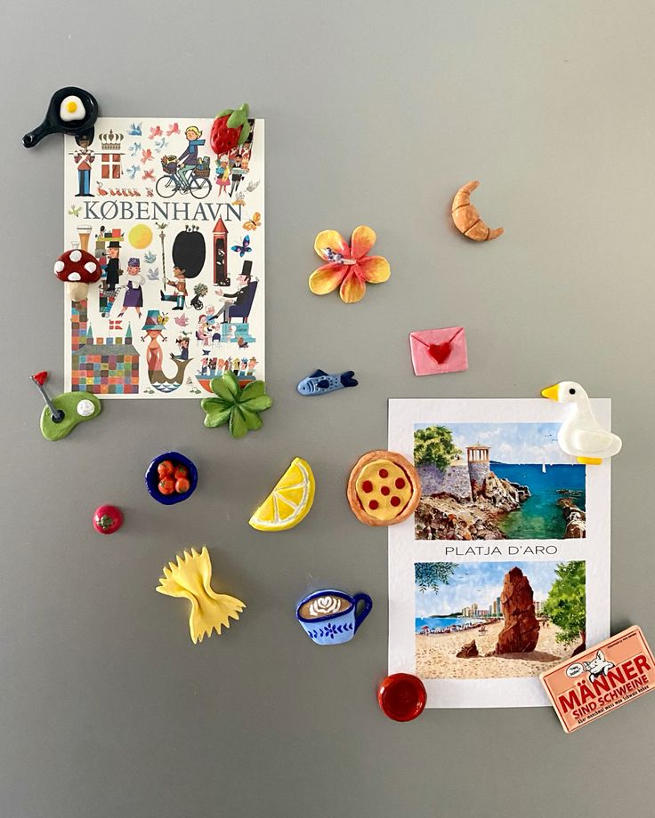 Colourful European-style clay magnets including a croissant, goose and lemon slice around travel postcards on a grey fridge