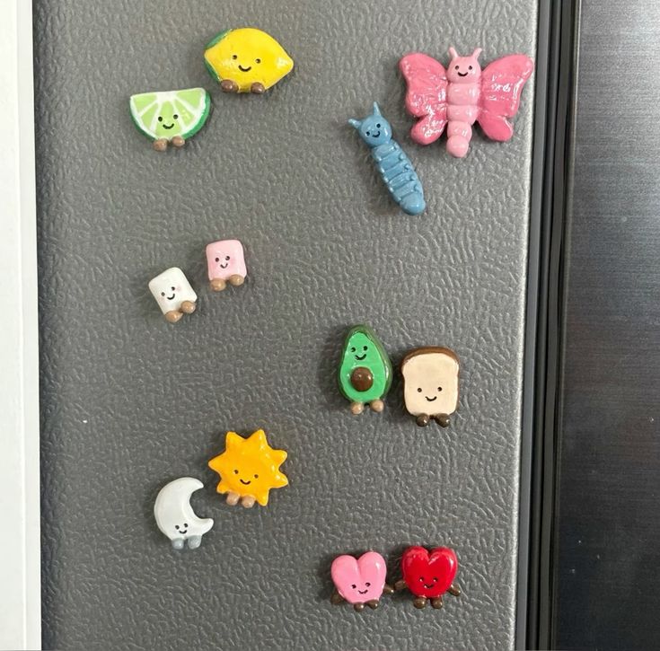 Smiling clay character magnets with faces and legs including a butterfly, caterpillar, sun and moon on a dark fridge