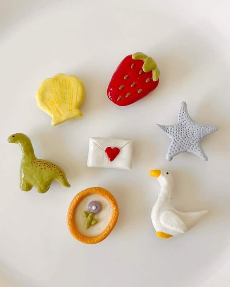Glossy glazed clay magnets including a dinosaur, goose, shell, starfish and heart envelope on a white plate
