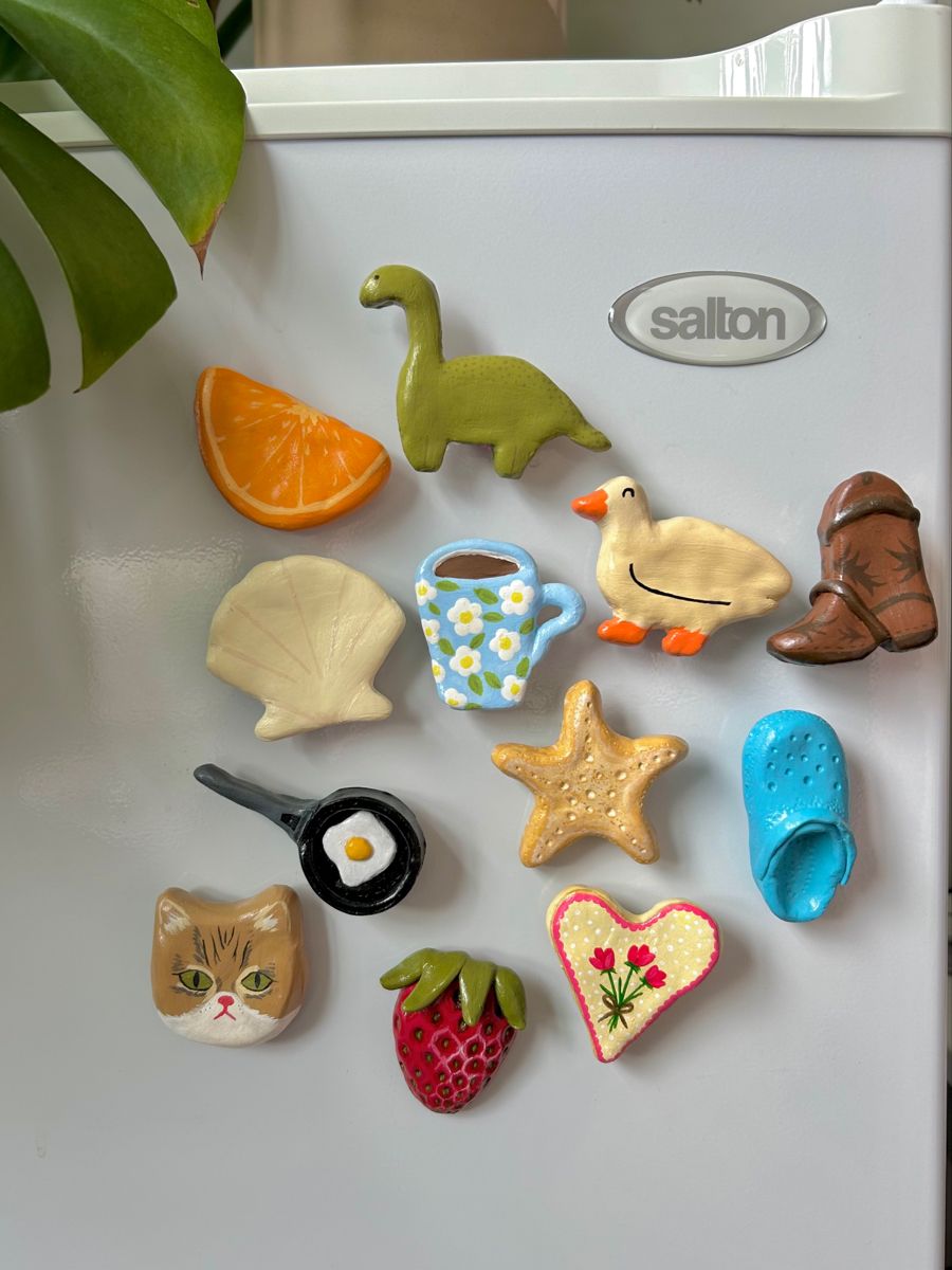 Matte hand-painted clay magnets including a cat, goose, cowboy boots, clog and strawberry on a white fridge