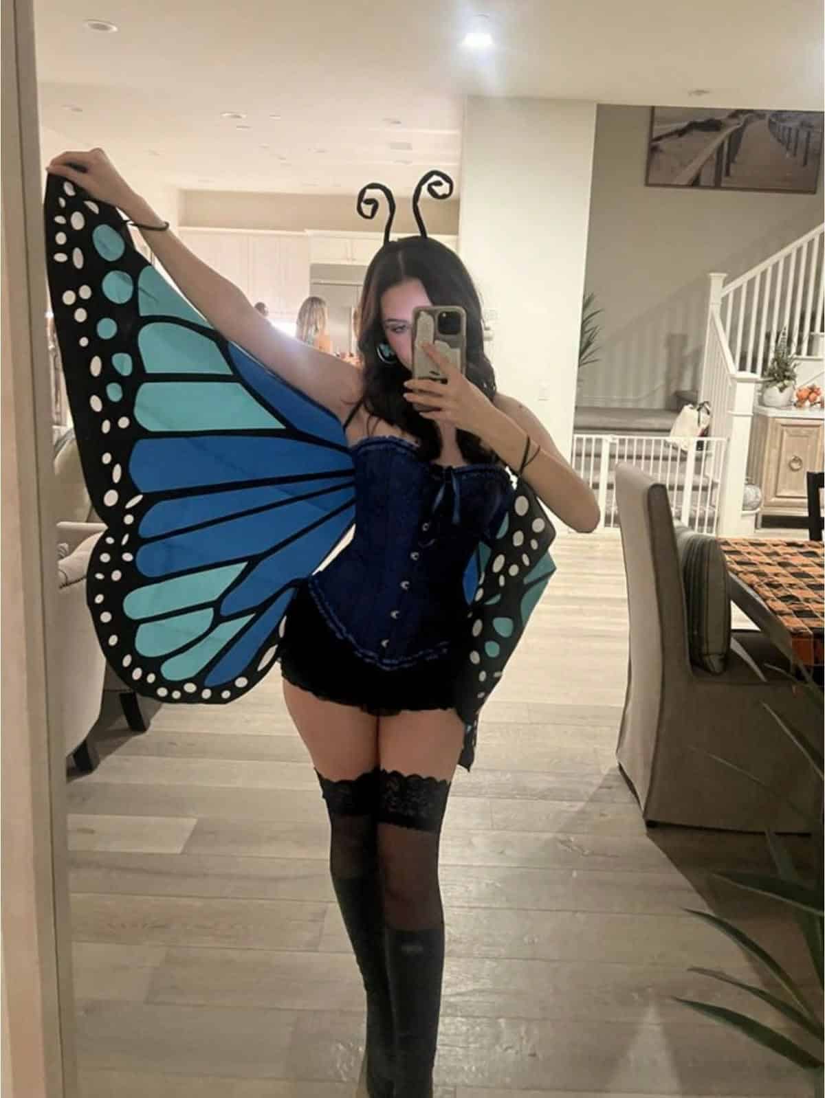 Person holding large blue monarch butterfly wings, wearing a corset, shorts and black stockings with butterfly antennae.