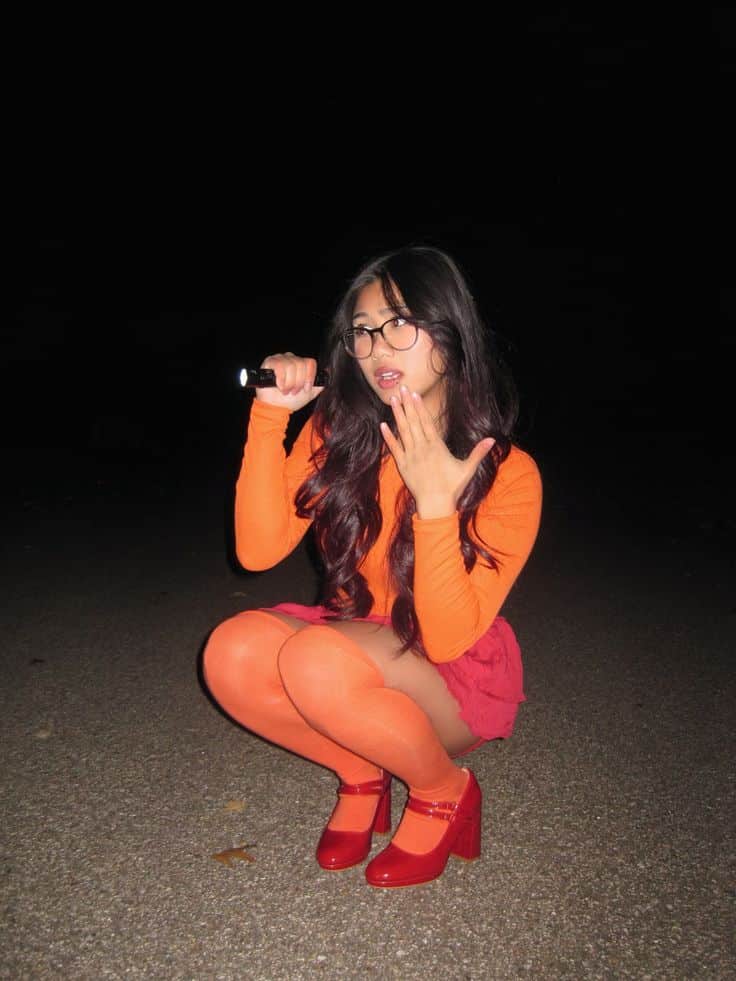 Person in an orange top, orange tights and glasses holding a torch, dressed as Velma from Scooby-Doo.
