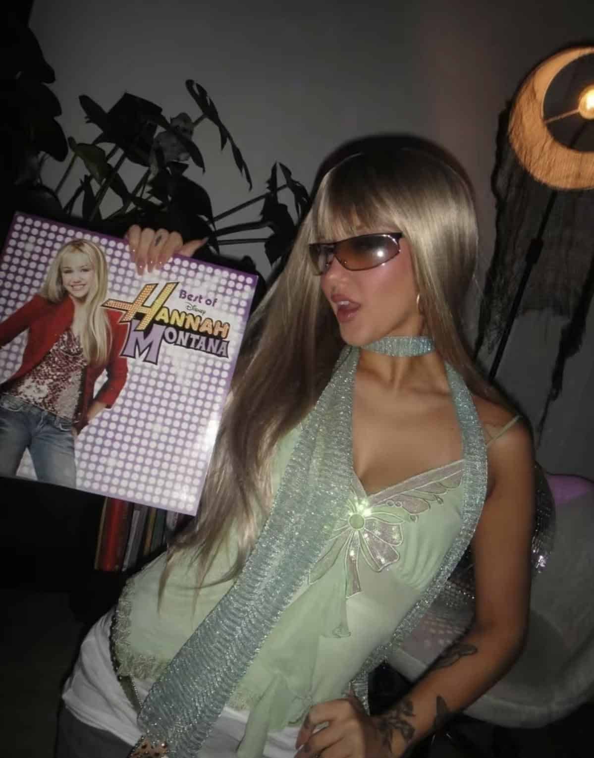 Person in a shimmery green halter and blonde wig with sunglasses, holding a Hannah Montana album.