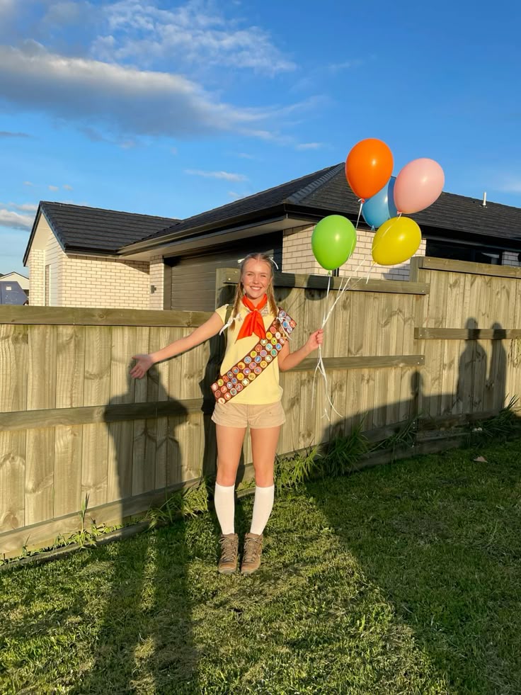 Person in a yellow shirt with a badge sash, red neckerchief and khaki shorts holding balloons, dressed as Russell from Up.