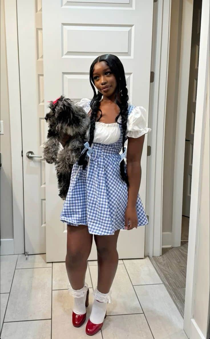 Person in a blue gingham pinafore and white top with braided hair and red shoes, holding a small dog, dressed as Dorothy.