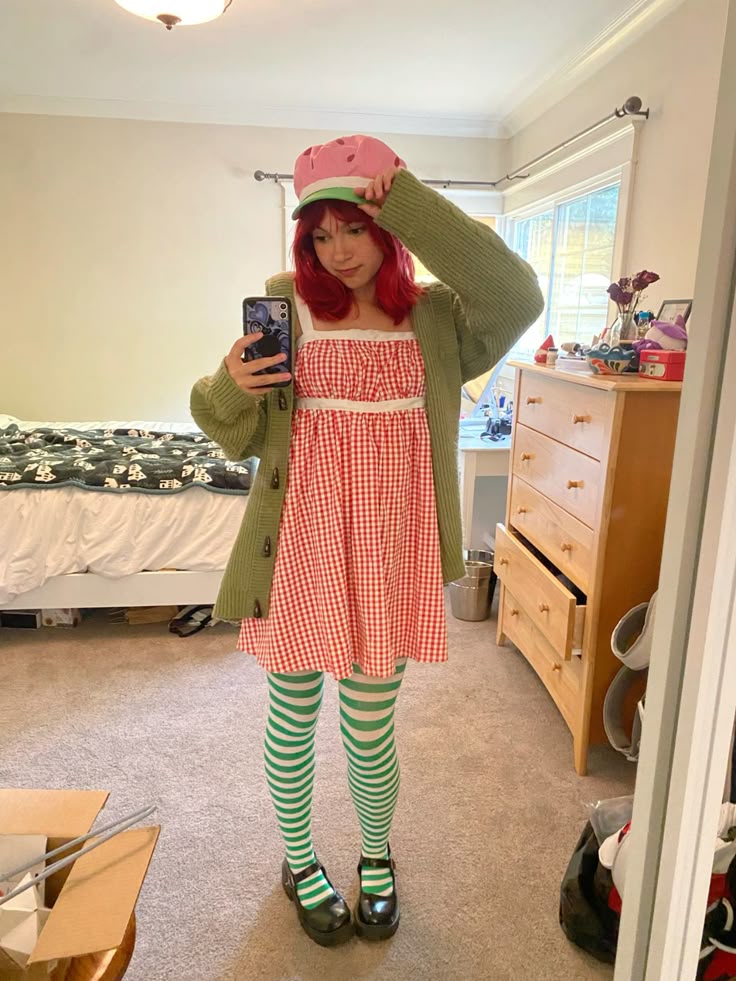 Person in a red gingham dress, green cardigan and green-striped tights with a fruit-themed cap, dressed as Strawberry Shortcake.