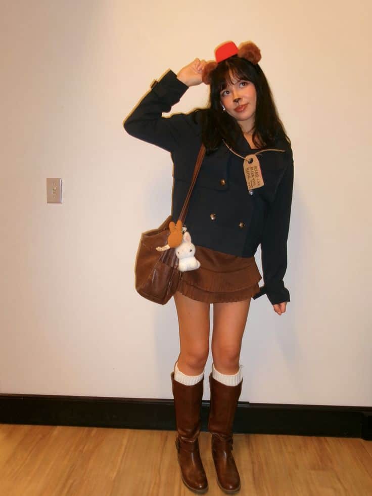 Person in a navy coat with bear ears and a luggage tag, corduroy skirt and knee boots, dressed as a Paddington-style bear.