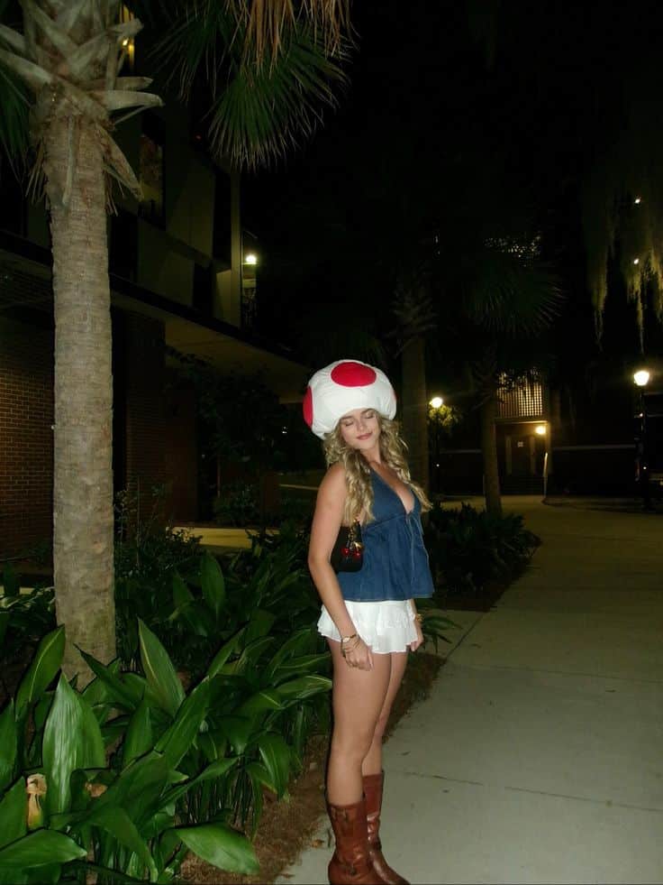 Person in a white mushroom cap with red spots, denim halter top and white ruffle skirt, dressed as Toad from Super Mario.