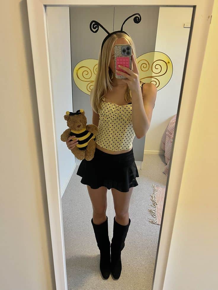 Person in a yellow polka-dot corset top with bee wings and antennae, holding a bee teddy.