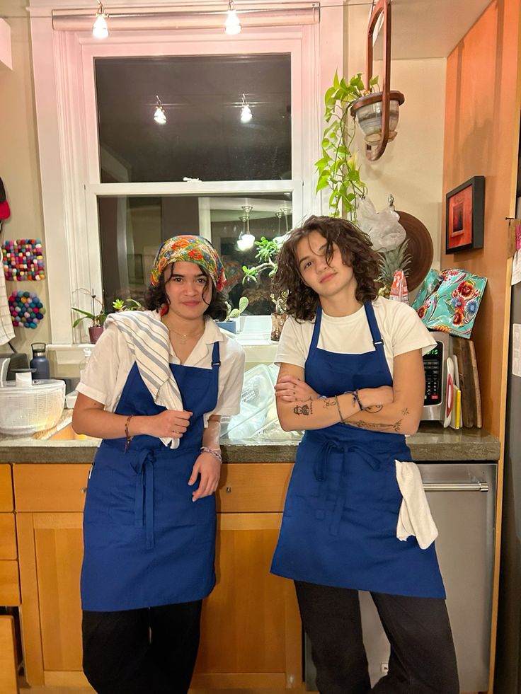 Two friends dressed as Carmy and Sydney from The Bear in matching blue aprons over white tees in a kitchen