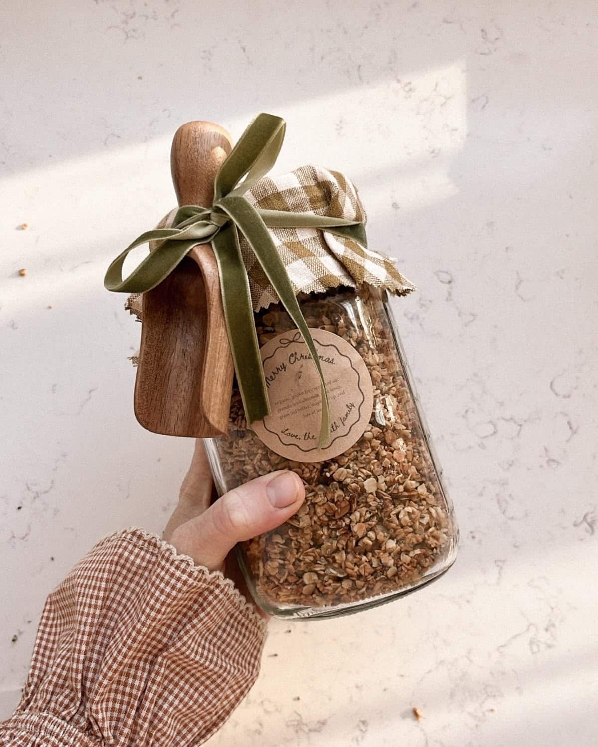 Large glass jar of homemade granola with a gingham fabric topper, green velvet ribbon and wooden scoop