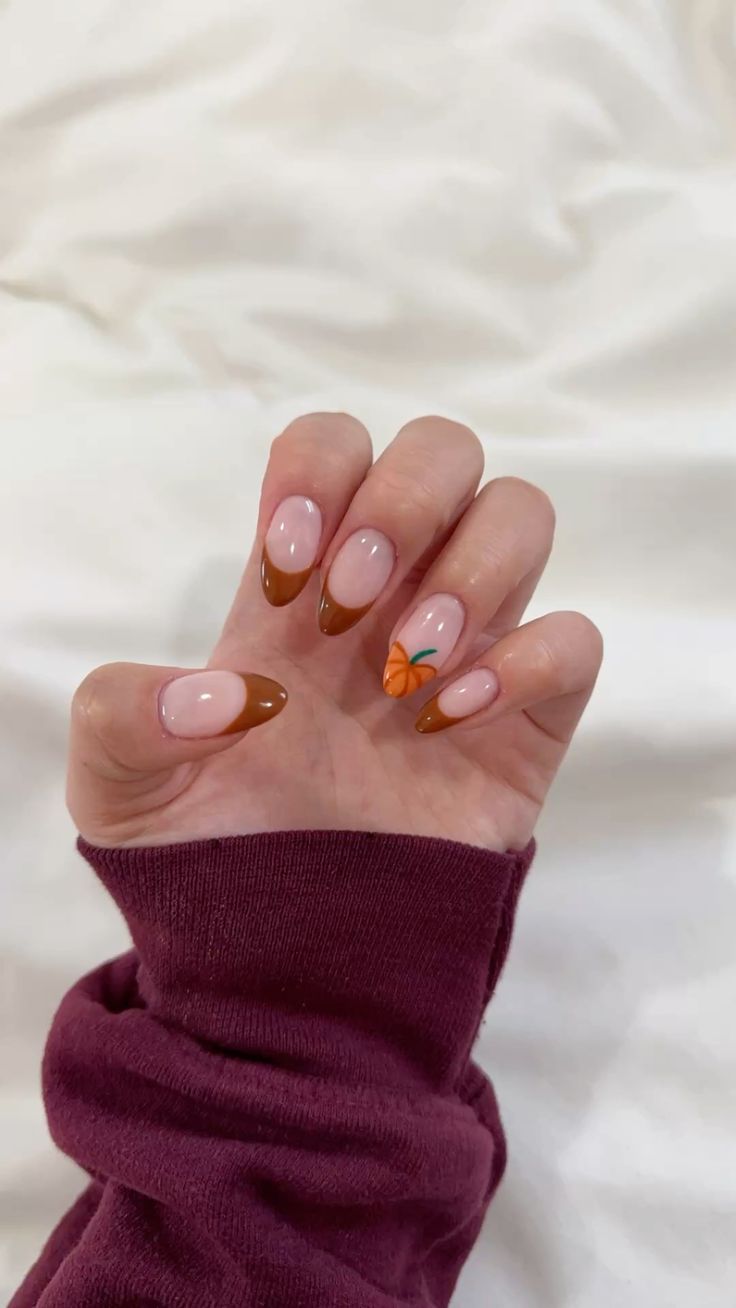 Almond nails with brown French tips on a sheer pink base and a hand-painted orange pumpkin accent nail